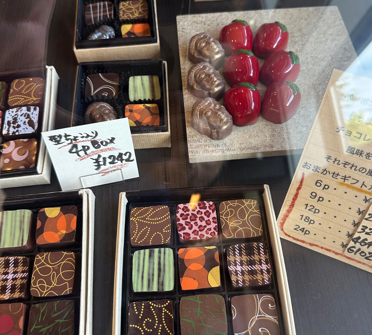 Assorted artisanal chocolates in open boxes with colorful patterns, plus glossy red bell‑pepper shaped chocolates on a stone display and a handwritten price sign nearby