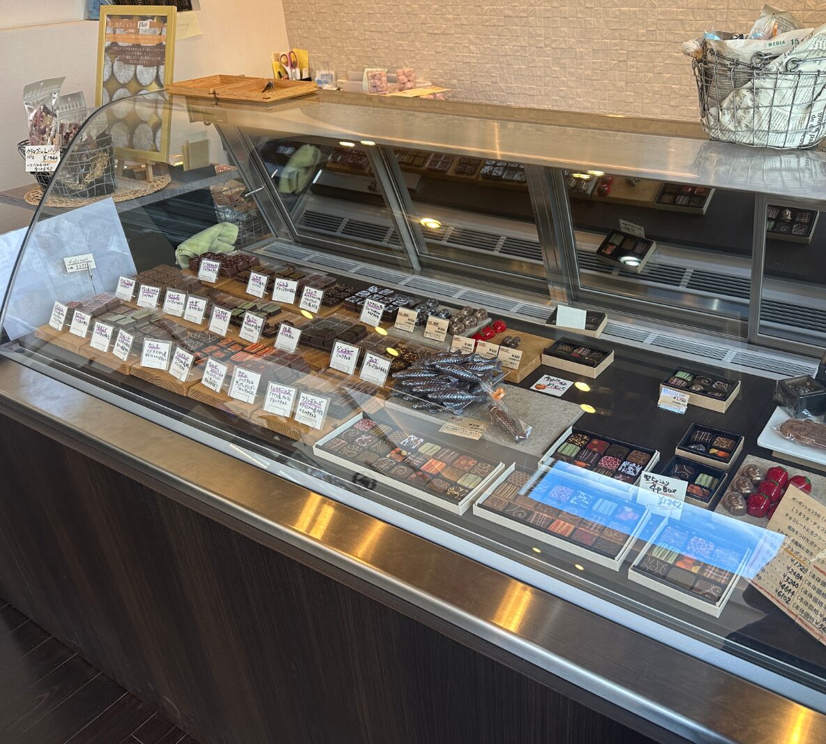 Glass-front chocolate display case with assorted truffles and pralines, small labels in Japanese indicating flavors, and a basket of wrapped chocolates on top left corner.