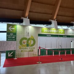 Exhibition booth area with green banners reading '2026 中部パック', red carpet, and a microphone stand in a convention hall.