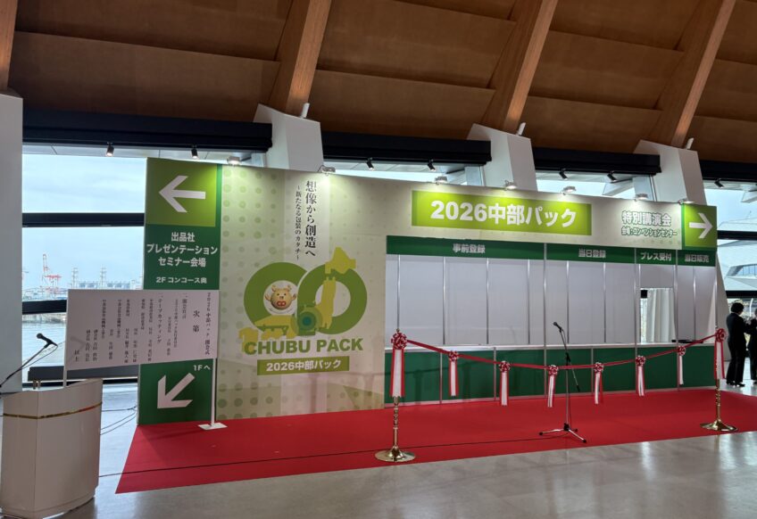 Exhibition booth area with green banners reading '2026 中部パック', red carpet, and a microphone stand in a convention hall.