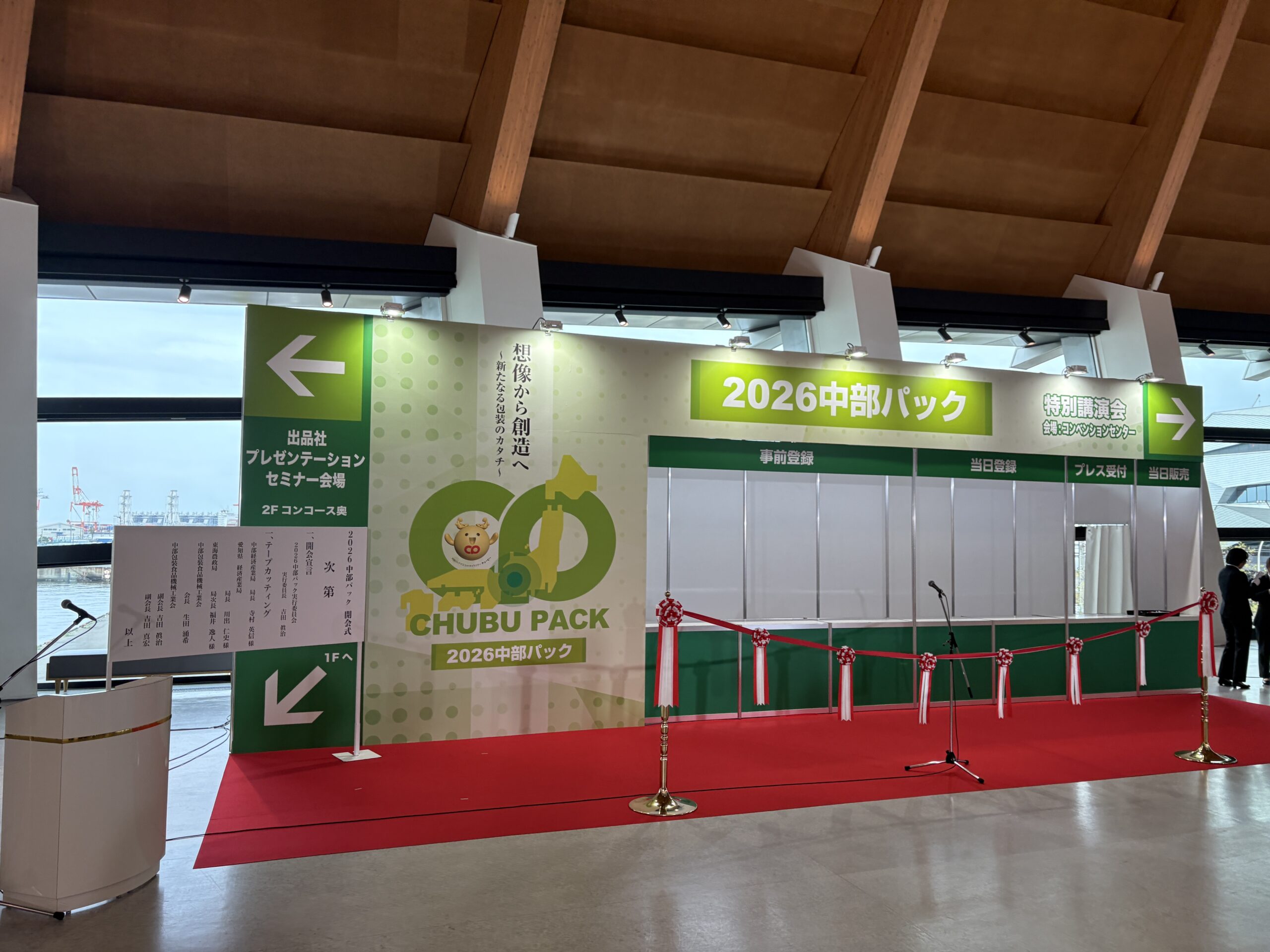 Exhibition booth area with green banners reading '2026 中部パック', red carpet, and a microphone stand in a convention hall.